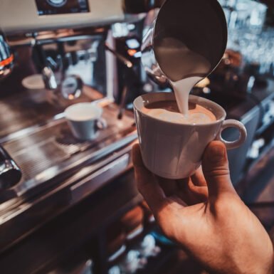 Diligent barista is prepairing fresh latte for customers for coffee break at caffeteria.